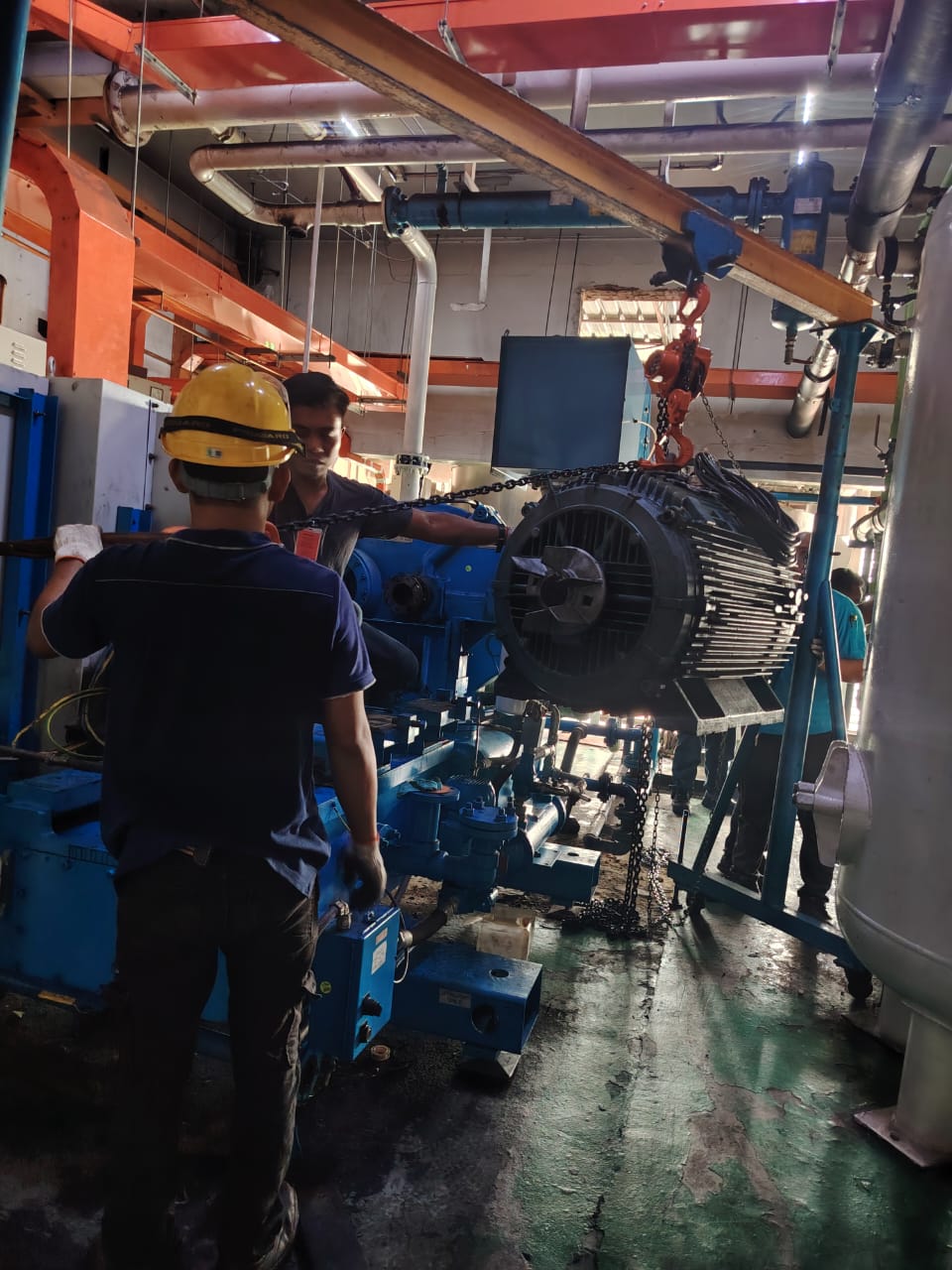 Technician in a hard hat guiding a large electric motor lifted on a chain hoist