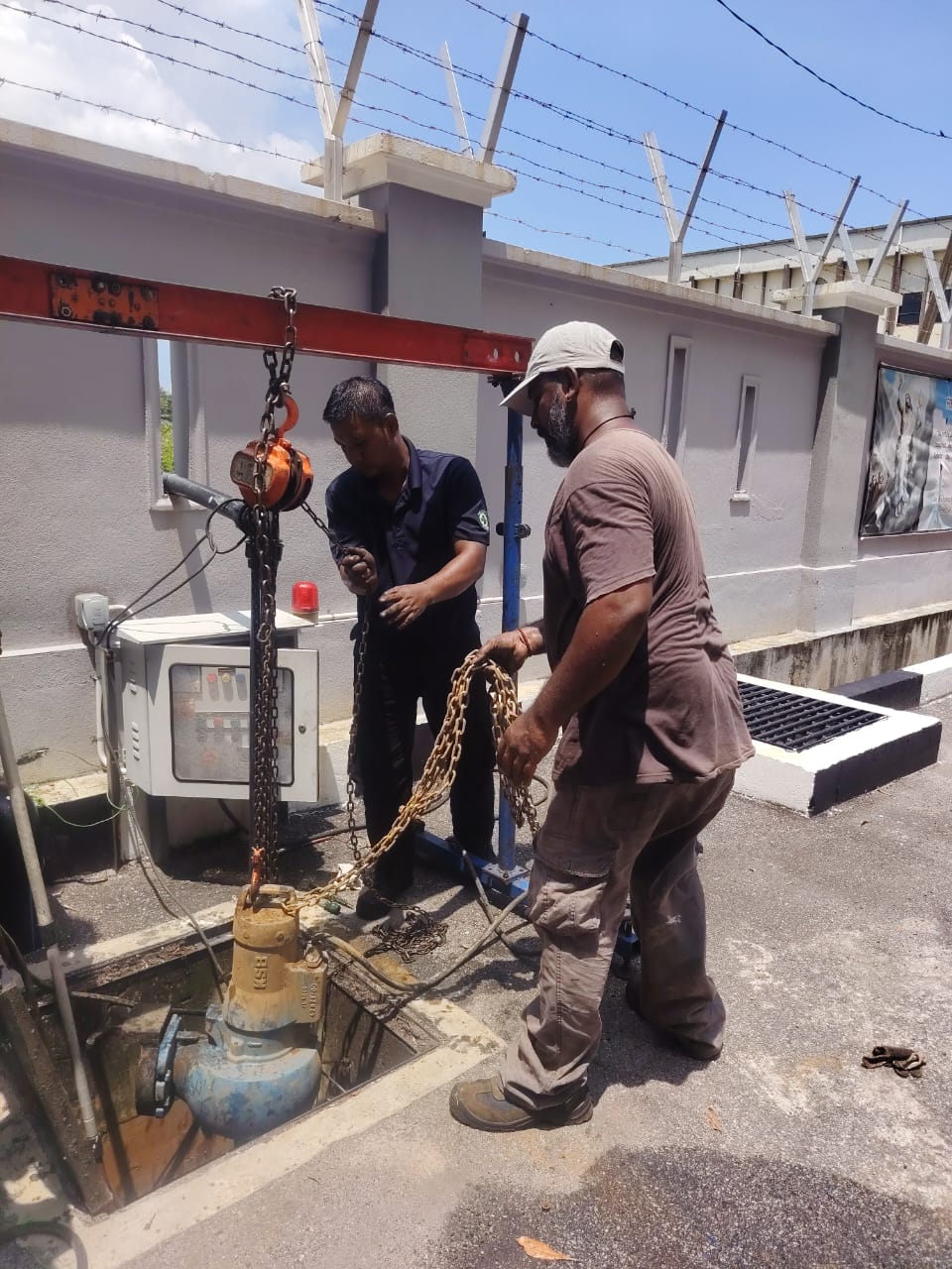 Two technicians lifting a submersible pump out of a sump with a chain hoist